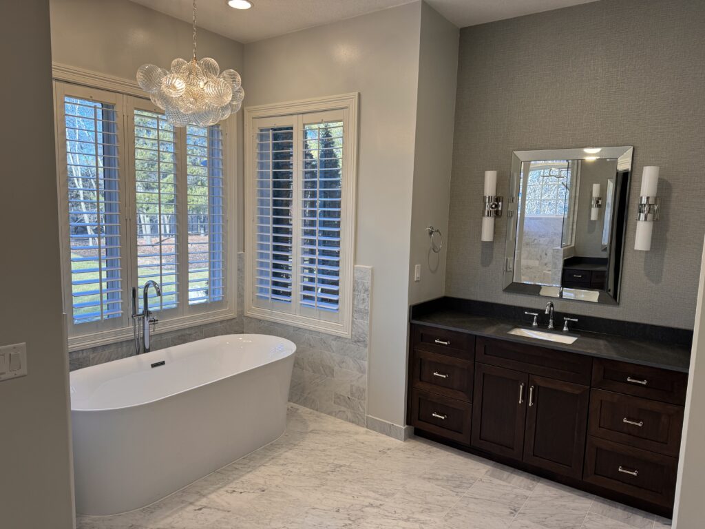 Modern bathroom remodel Edina MN with a freestanding white tub by large windows with white shutters, marble floor, dark wood vanity with sink, wall-mounted mirror, and contemporary light fixtures.
