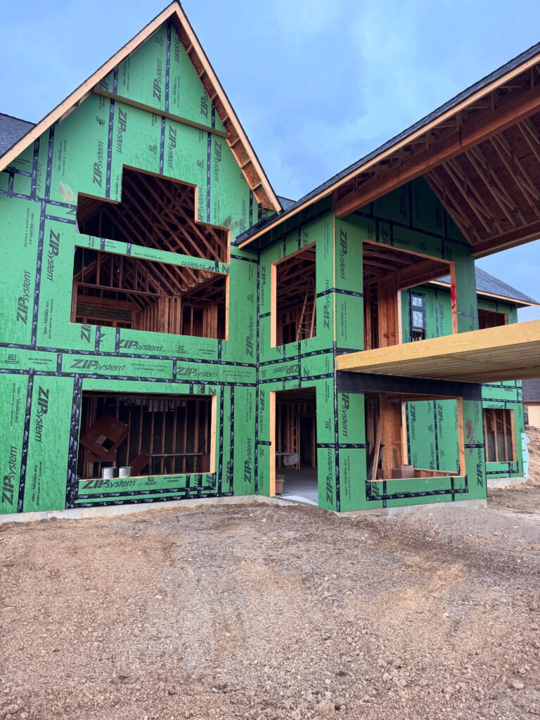 A two-story house under construction by a custom home builder in Lakeville, MN, featuring exposed wooden framing, large window openings, and using energy efficient zip panel sheeting on green sheathing panels. Unfinished dirt ground is seen in the foreground.