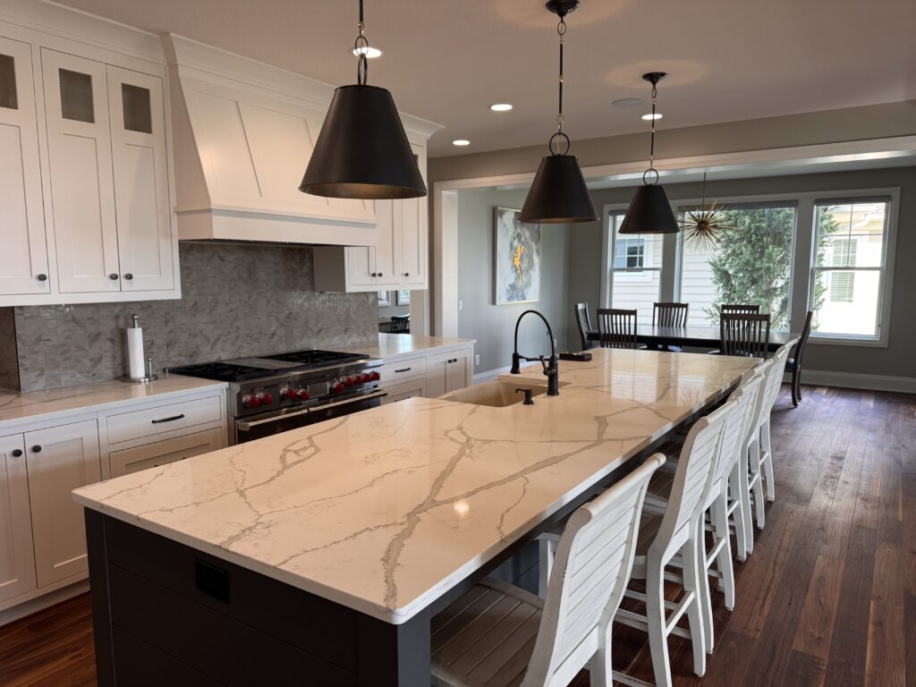 A modern kitchen by Lakeville-MN-Custom-Home-Builder features a large white marble island, black pendant lights, white cabinets, a built-in stove, and a dining area with a table and chairs near large windows.
